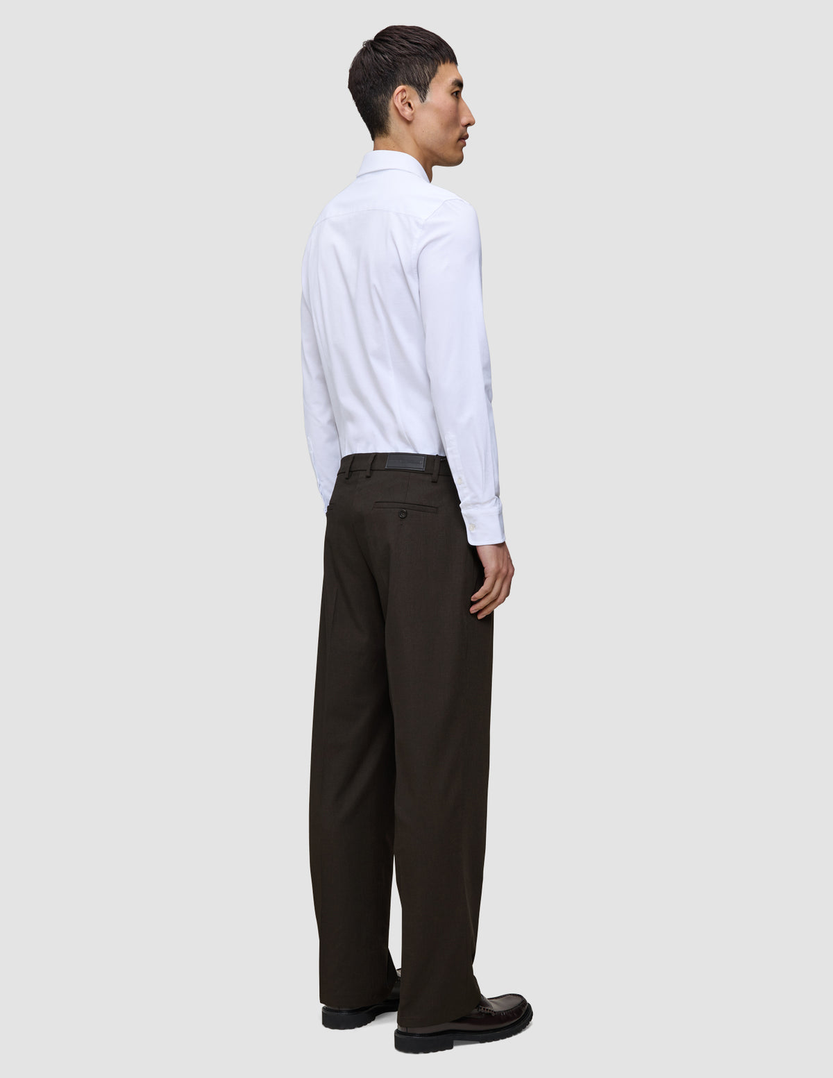 En person klädd i en vit skjorta med knappar och bruna Motion Suit-byxor i modellen Relaxed Cognac med 360°-stretch.