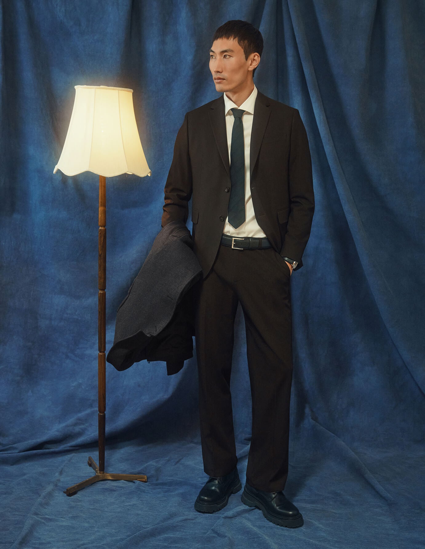 En man klädd i byxor av modellen Motion Suit Pants Relaxed Cognac, en vit skjorta och en slips står bredvid en golvlampa.