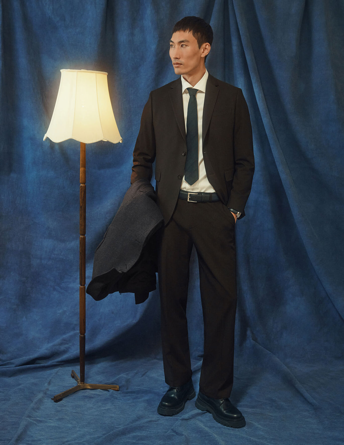 En man klädd i byxor av modellen Motion Suit Pants Relaxed Cognac, en vit skjorta och en slips står bredvid en golvlampa.