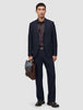 En man klädd i mörkblå byxor i modell Midnight Blue Motion Suit med avslappnad passform, en mörk skjorta och bruna loafers håller i en läderportfölj.