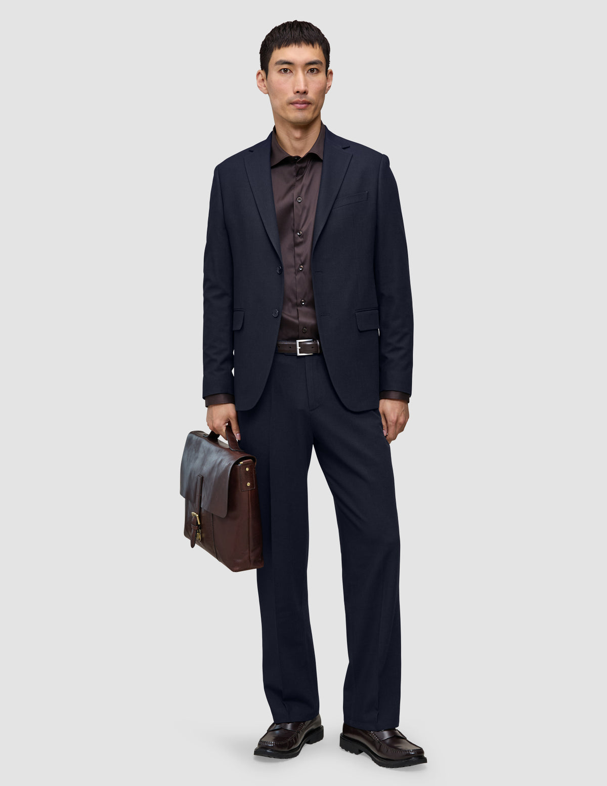 En man klädd i mörkblå byxor i modell Midnight Blue Motion Suit med avslappnad passform, en mörk skjorta och bruna loafers håller i en läderportfölj.