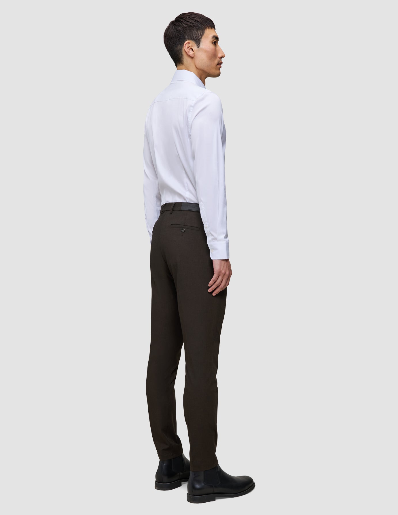 En man i en vit skjorta och mörka Motion Suit-byxor i modellen Slim Cognac står i profil.
