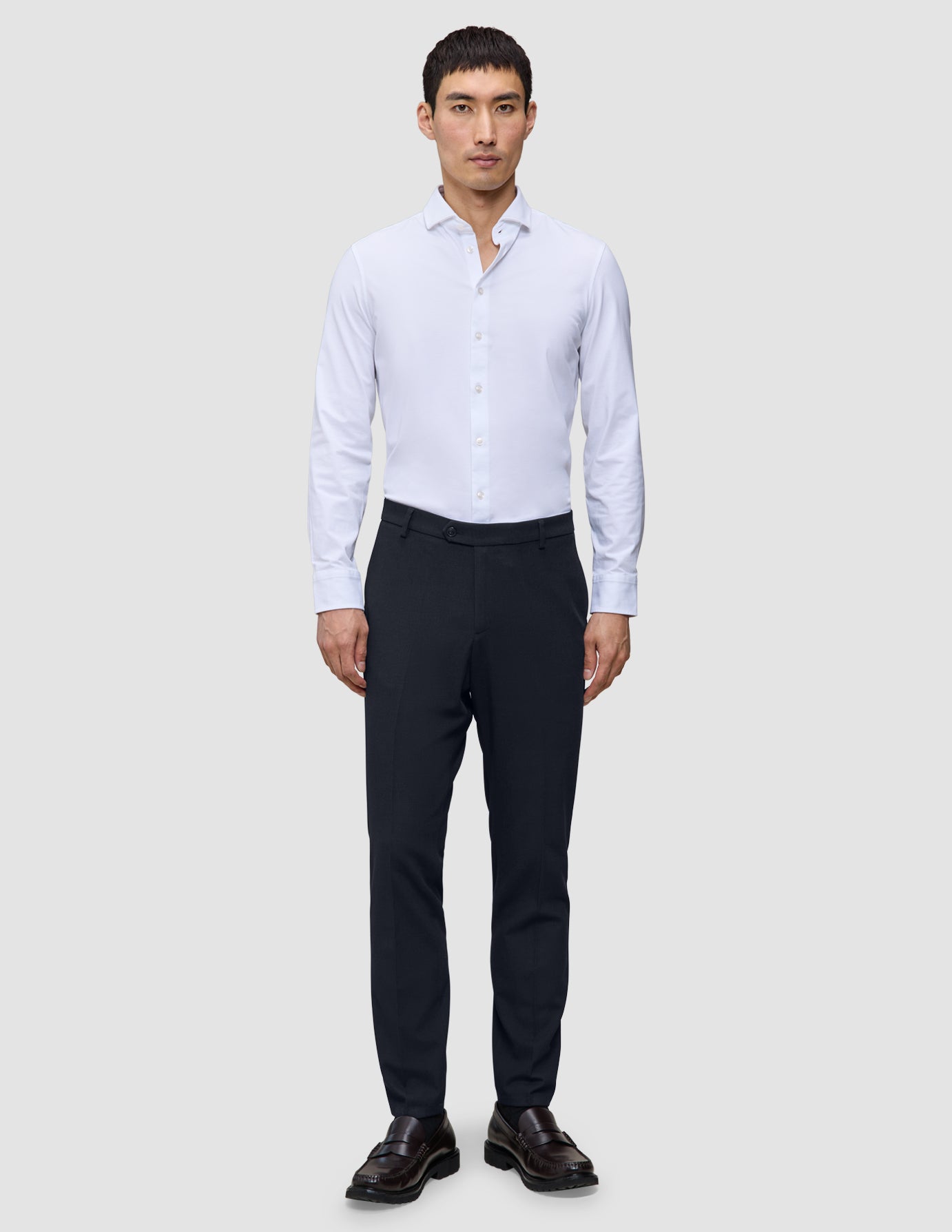 En man bär en vit skjorta med knappar, Motion Suit-byxor i smal passform i midnattblått och bruna loafers.