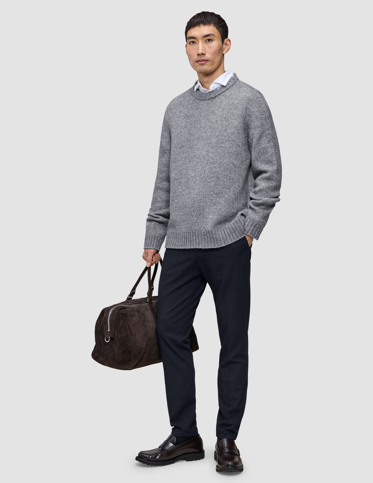En man klädd i en grå tröja, vit skjorta och ett par midnattsblå Motion Suit-byxor i slim fit håller i en brun resväska.