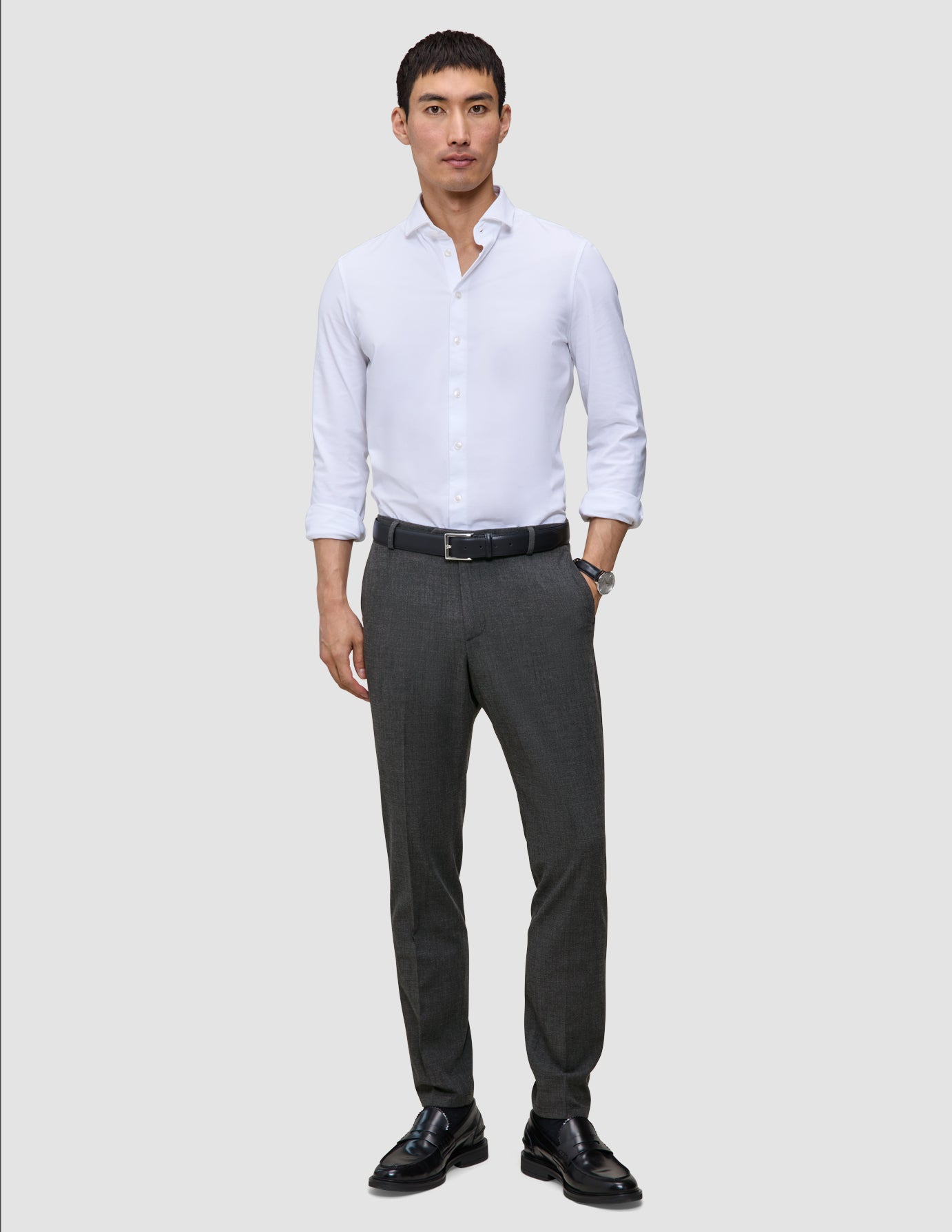 En man i vit skjorta och Motion Suit-byxor i modell Slim i färgen Pepper Grey, med svart bälte och loafers i skandinavisk stil.