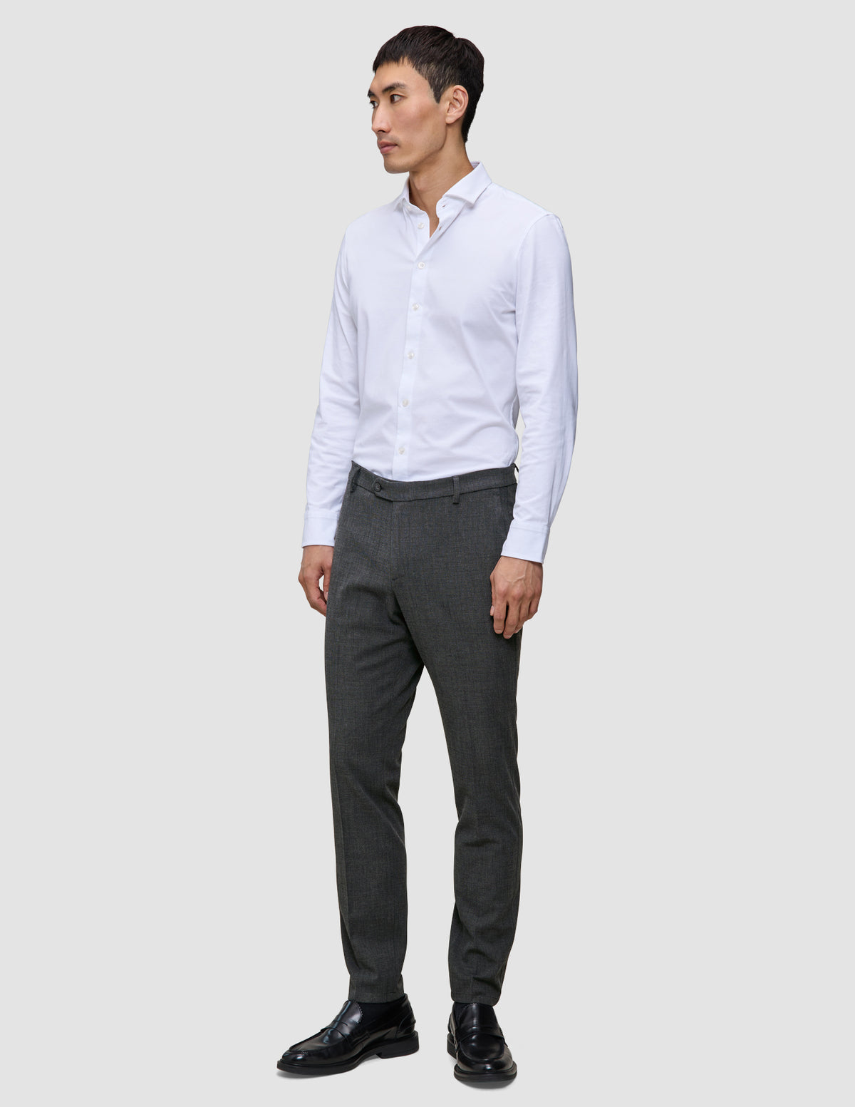 En man klädd i en vit skjorta med knappar, byxor av modellen Motion Suit Pants Slim Pepper Grey och svarta kostymskor.