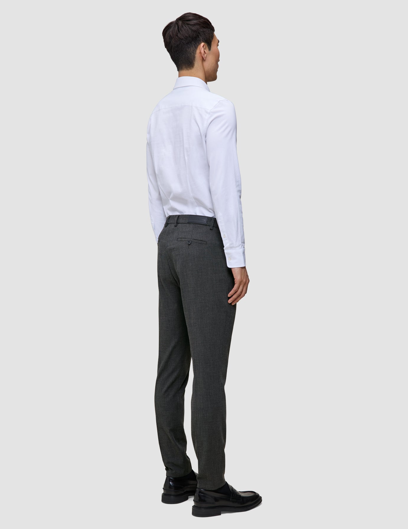 En person sedd bakifrån, klädd i en vit skjorta med knappar, Motion Suit-byxor i modell Slim i färgen Pepper Grey samt svarta kostymskor.