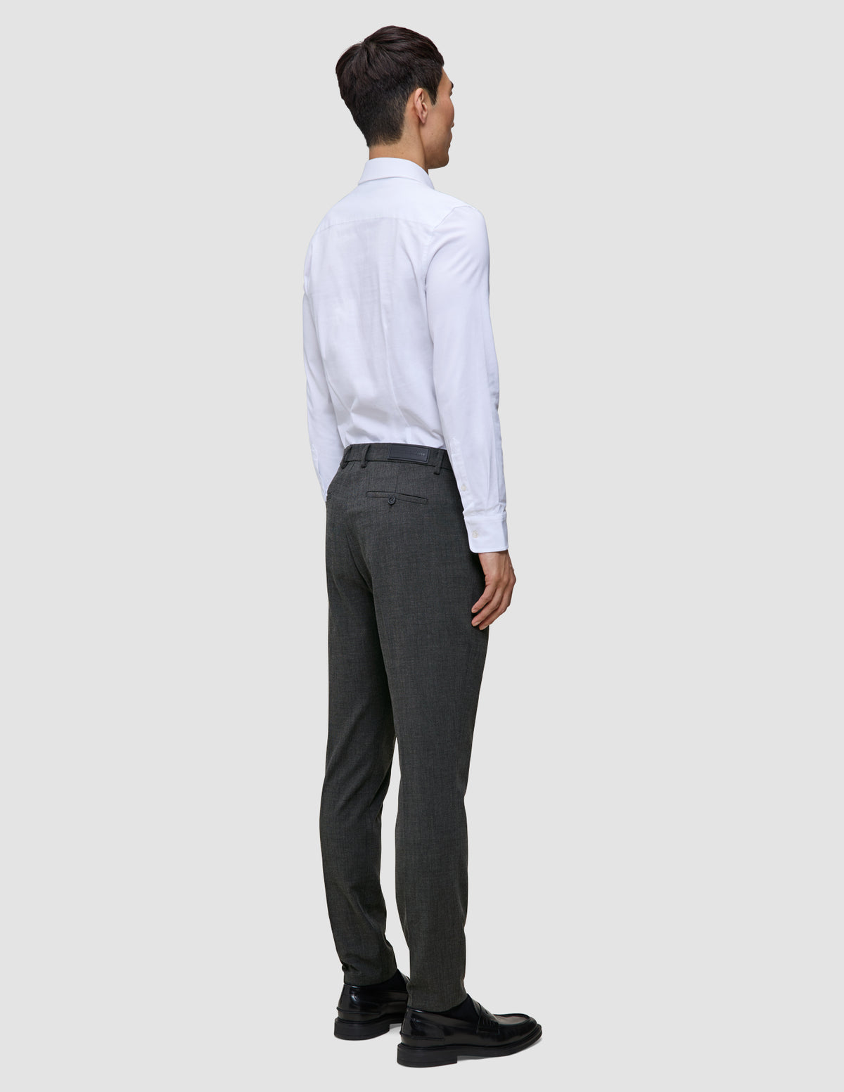 En person sedd bakifrån, klädd i en vit skjorta med knappar, Motion Suit-byxor i modell Slim i färgen Pepper Grey samt svarta kostymskor.