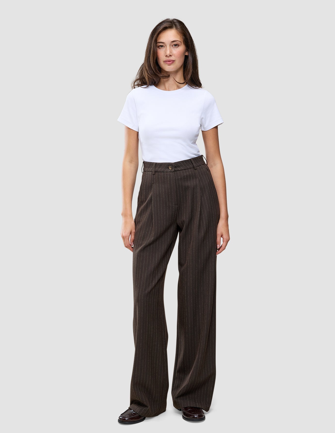 En person bär en vit t-shirt och byxorna Serene Pants Wide Walnut Herringbone: bruna byxor med hög midja och vida ben.