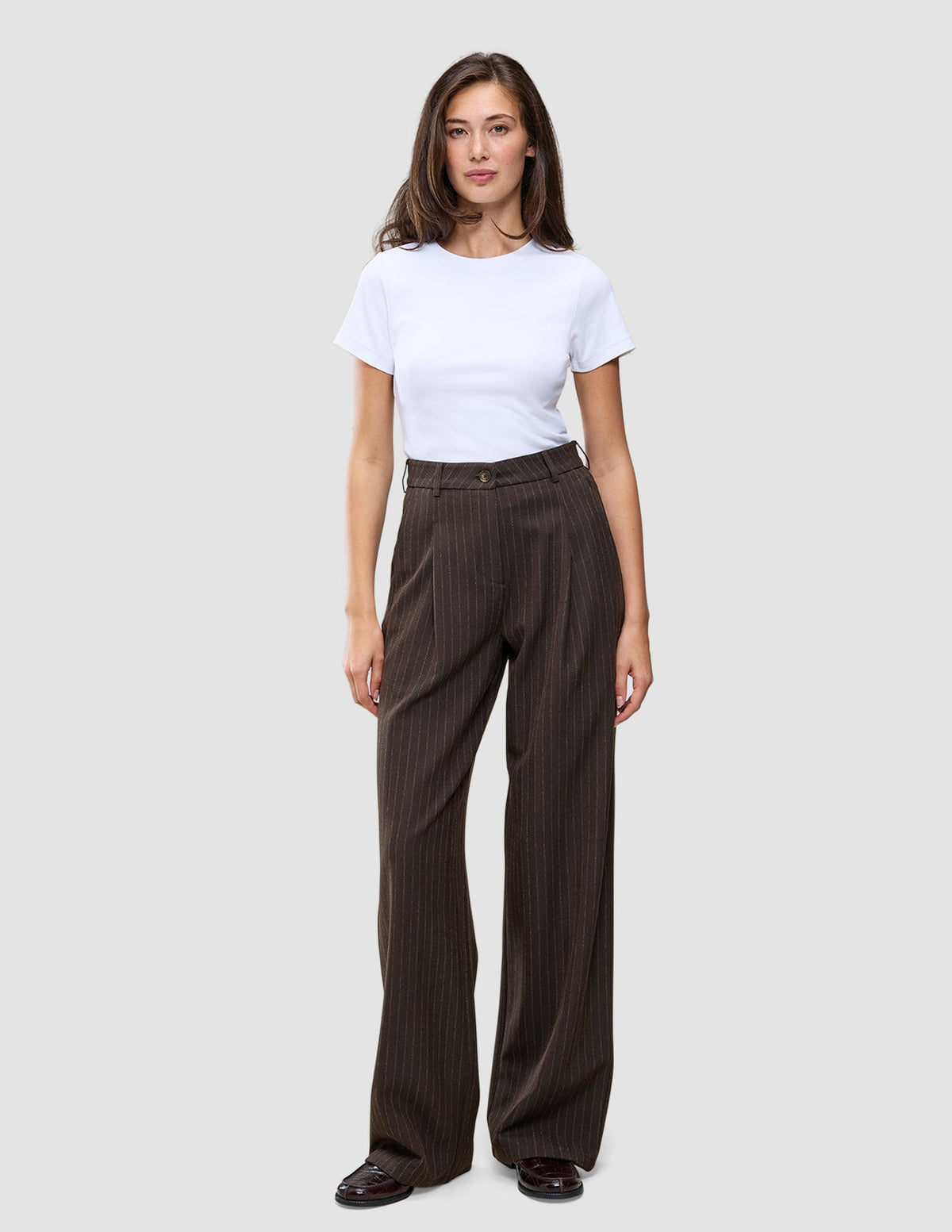 En person bär en vit t-shirt och byxorna Serene Pants Wide Walnut Herringbone: bruna byxor med hög midja och vida ben.