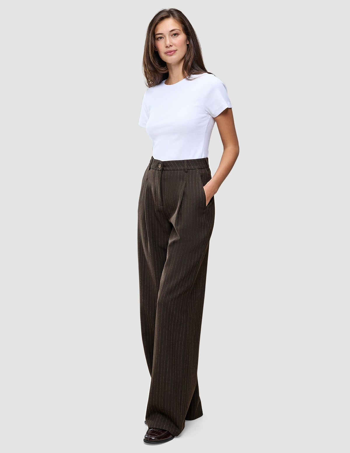 En kvinna i en vit t-shirt bär mörkbruna byxor med vida ben, modellen Serene Pants Wide Walnut Herringbone, med handen i fickan.