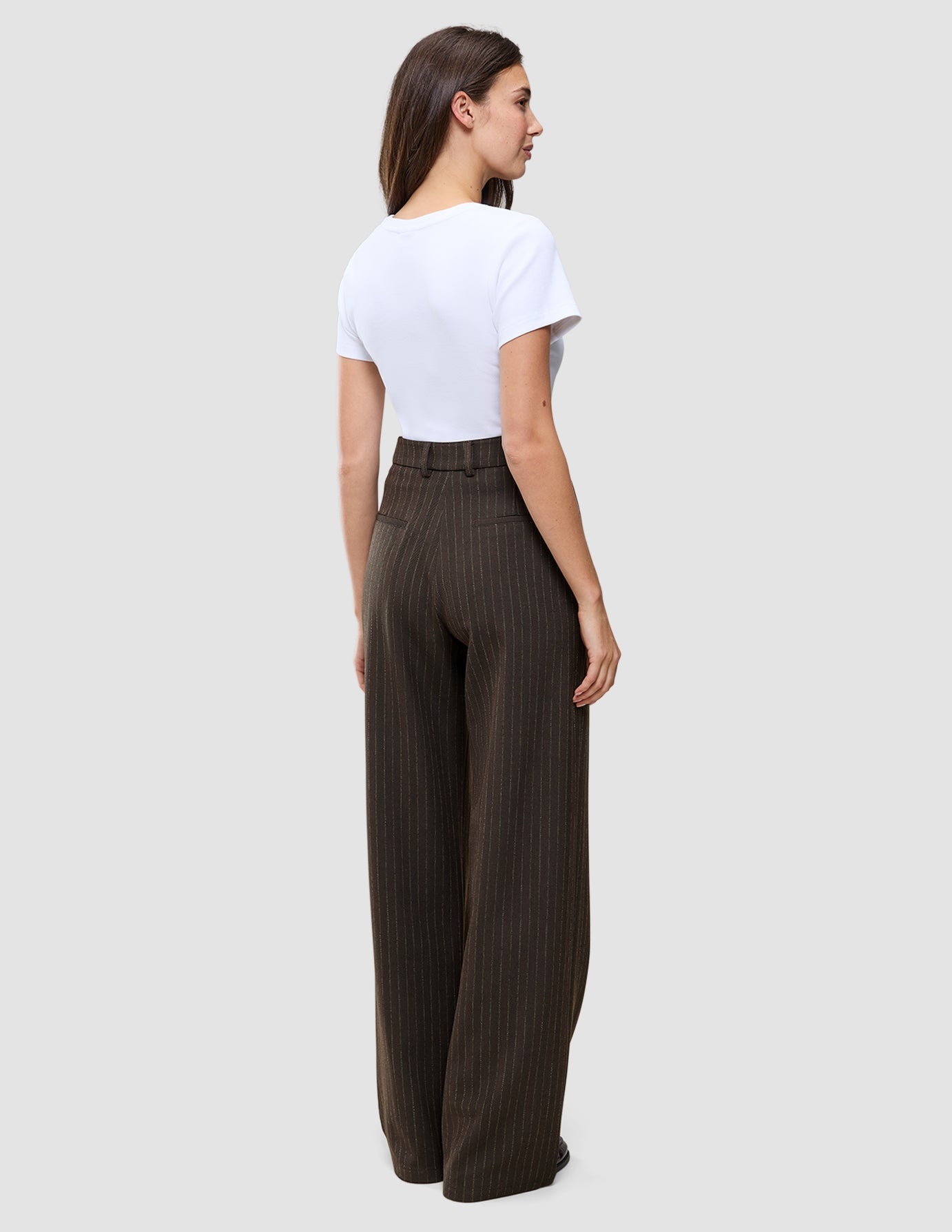 Kvinna i en vit t-shirt och mörkbruna, högmidjade, randiga byxor med vida ben av modellen Serene Pants Wide Walnut Herringbone.