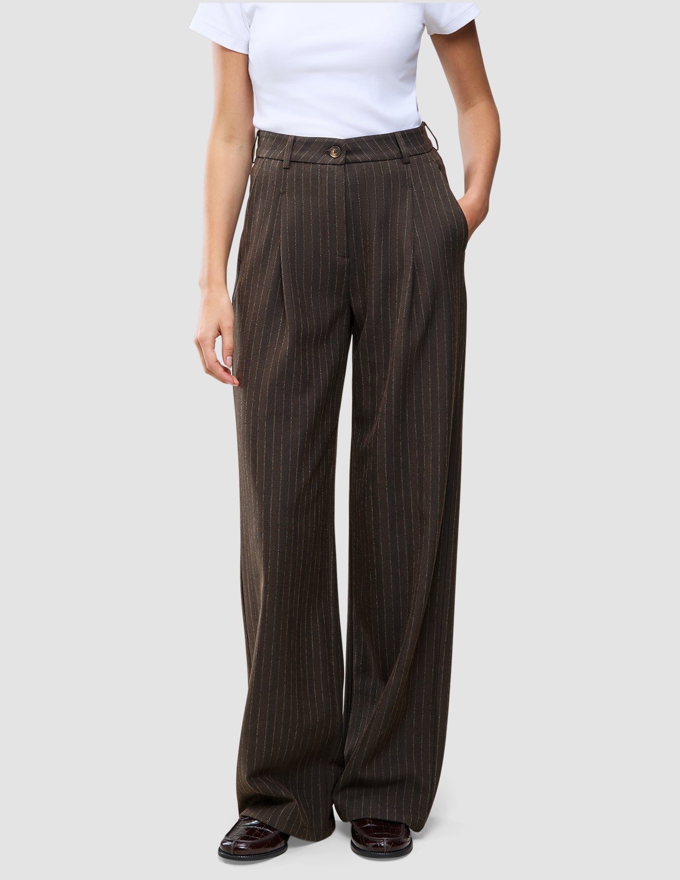 En person som bär ett par bruna Serene Pants Wide Walnut Herringbone-byxor med hög midja och vida ben, tillsammans med en vit t-shirt.