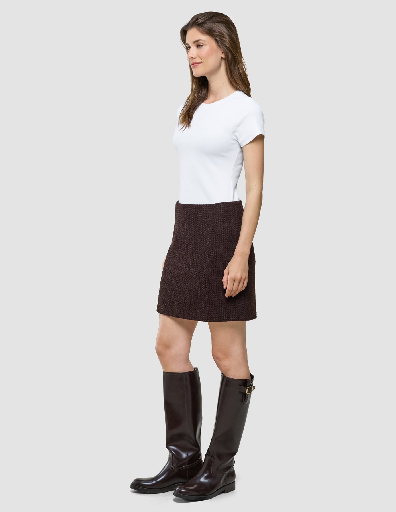 En kvinna klädd i en vit t-shirt, en kort kjol från Women Sorae i färgen Espresso och knähöga stövlar.