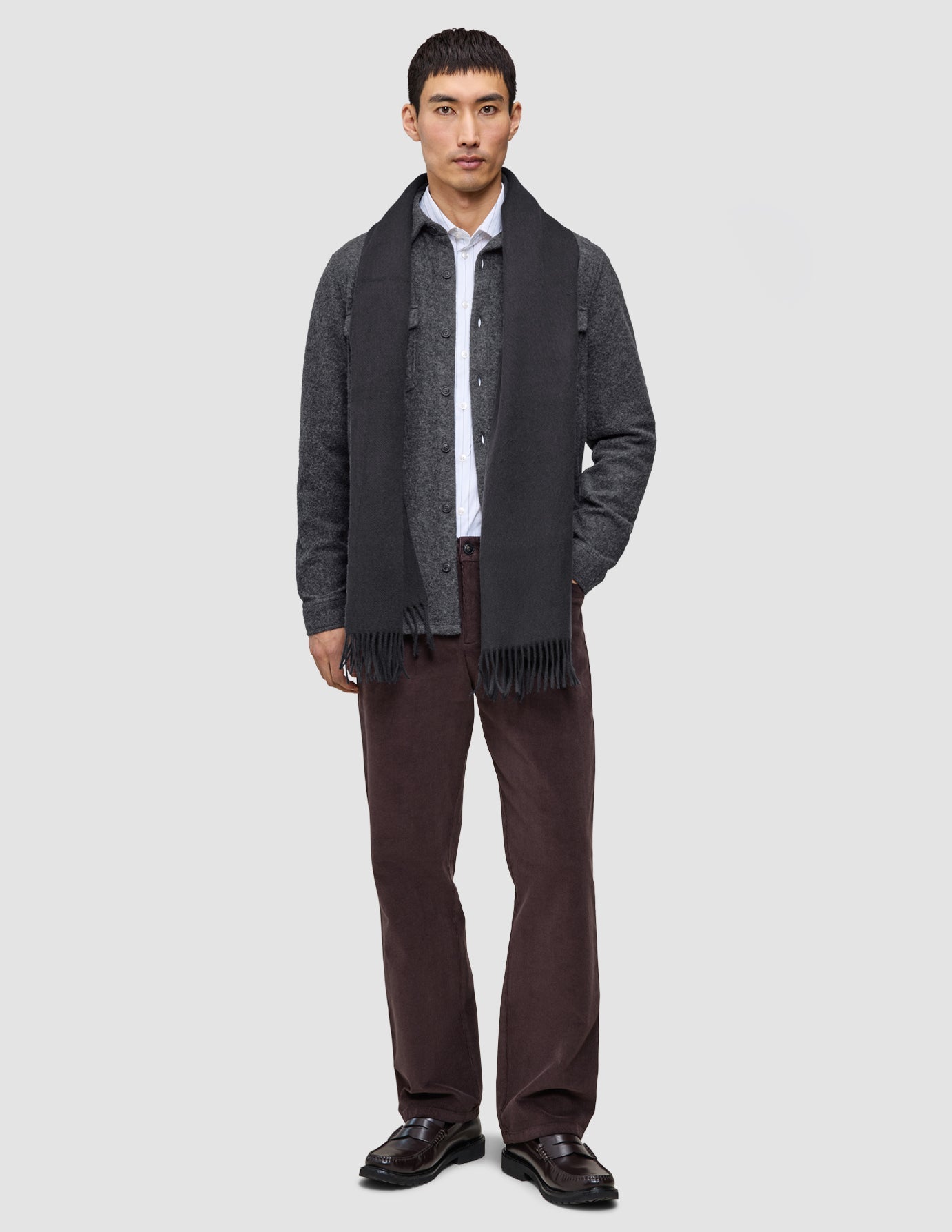 En man bär en avslappnad och stilfull övertröja över en vit skjorta, tillsammans med en mörk halsduk, bruna byxor och loafers.