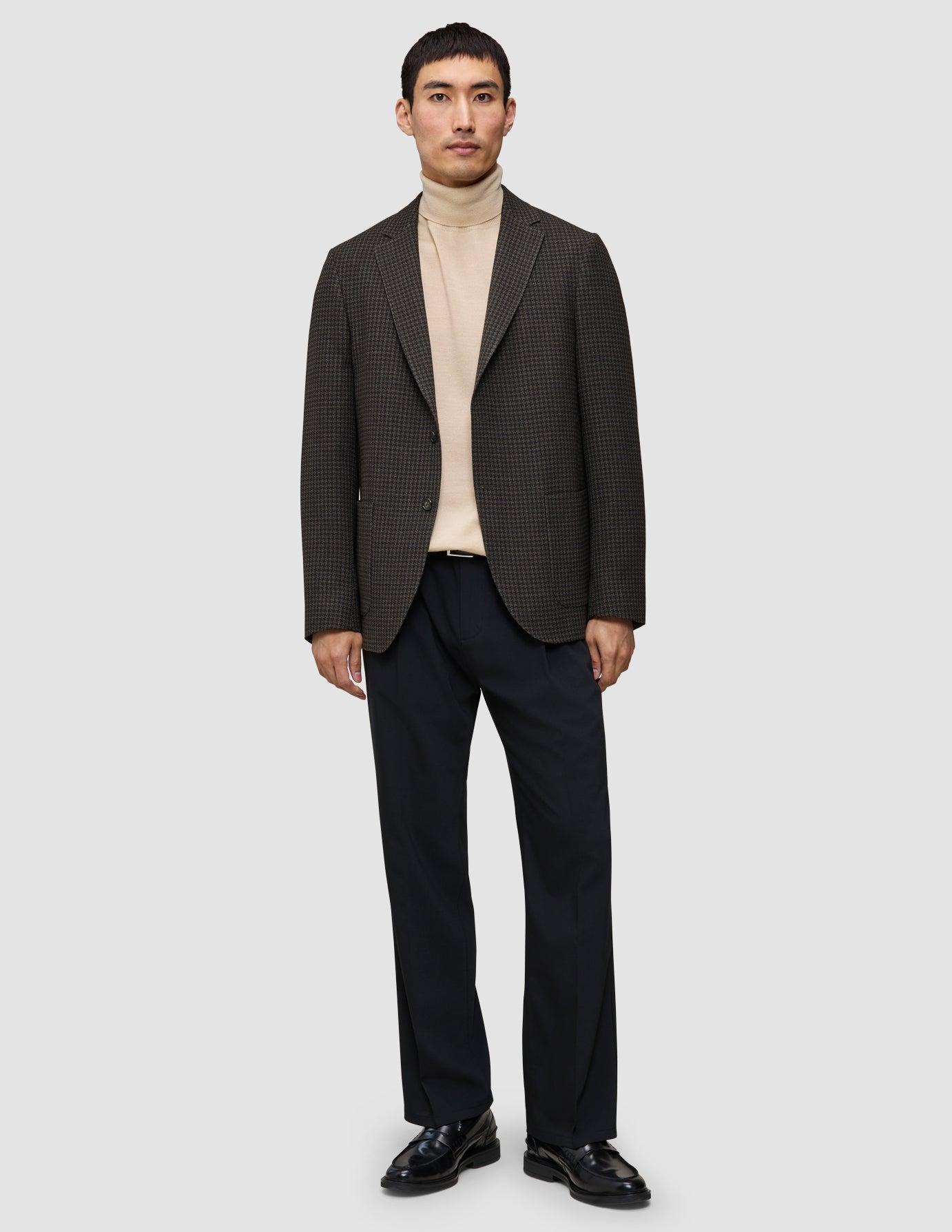 En man klädd i en kombination av stickat och kavaj: en mönstrad ullkavaj över en merino-tröja med rund hals, svarta byxor och svarta loafers.