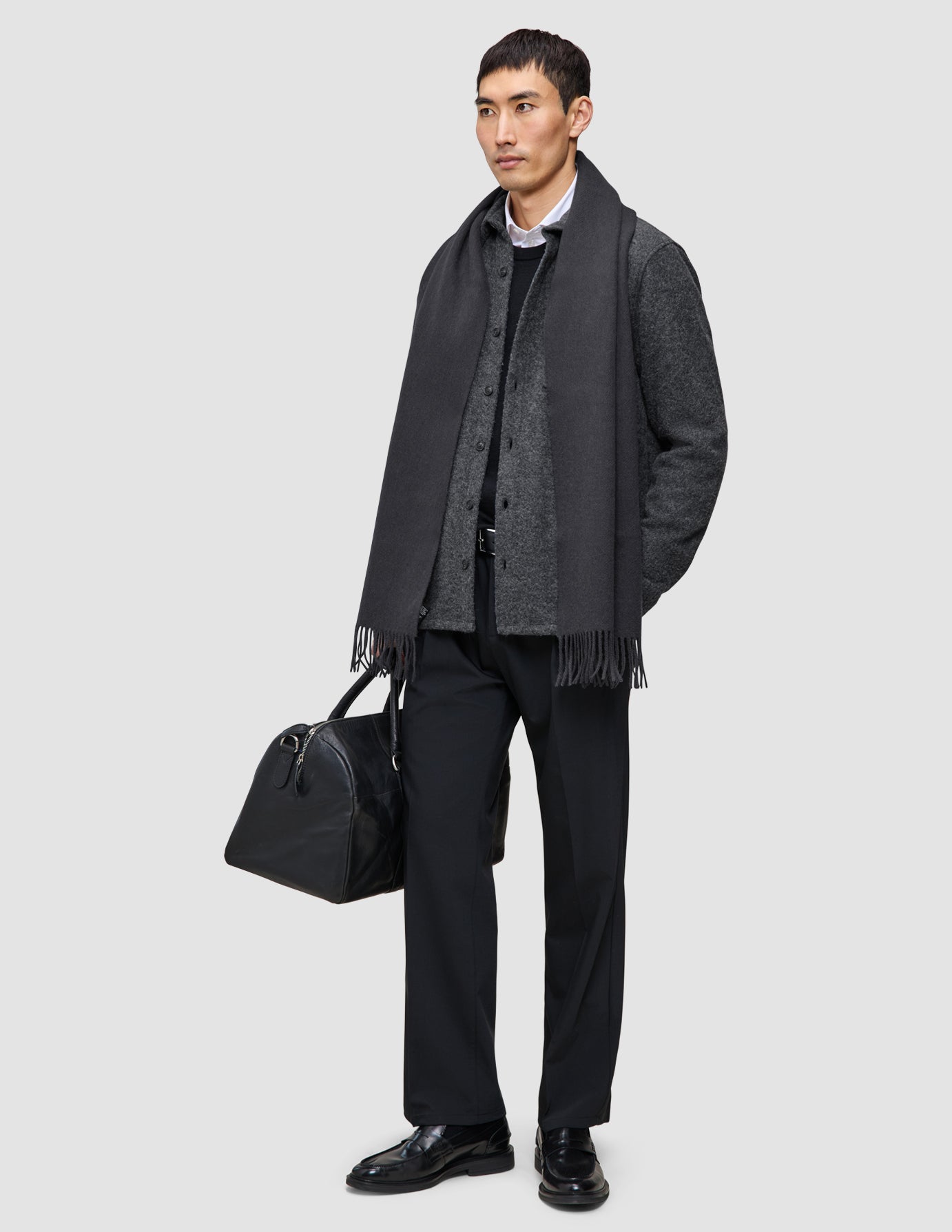 En man klädd i en väl sammansatt outfit: en grå halsduk, en övertröja i ull, byxor och loafers, som håller i en svart resväska.