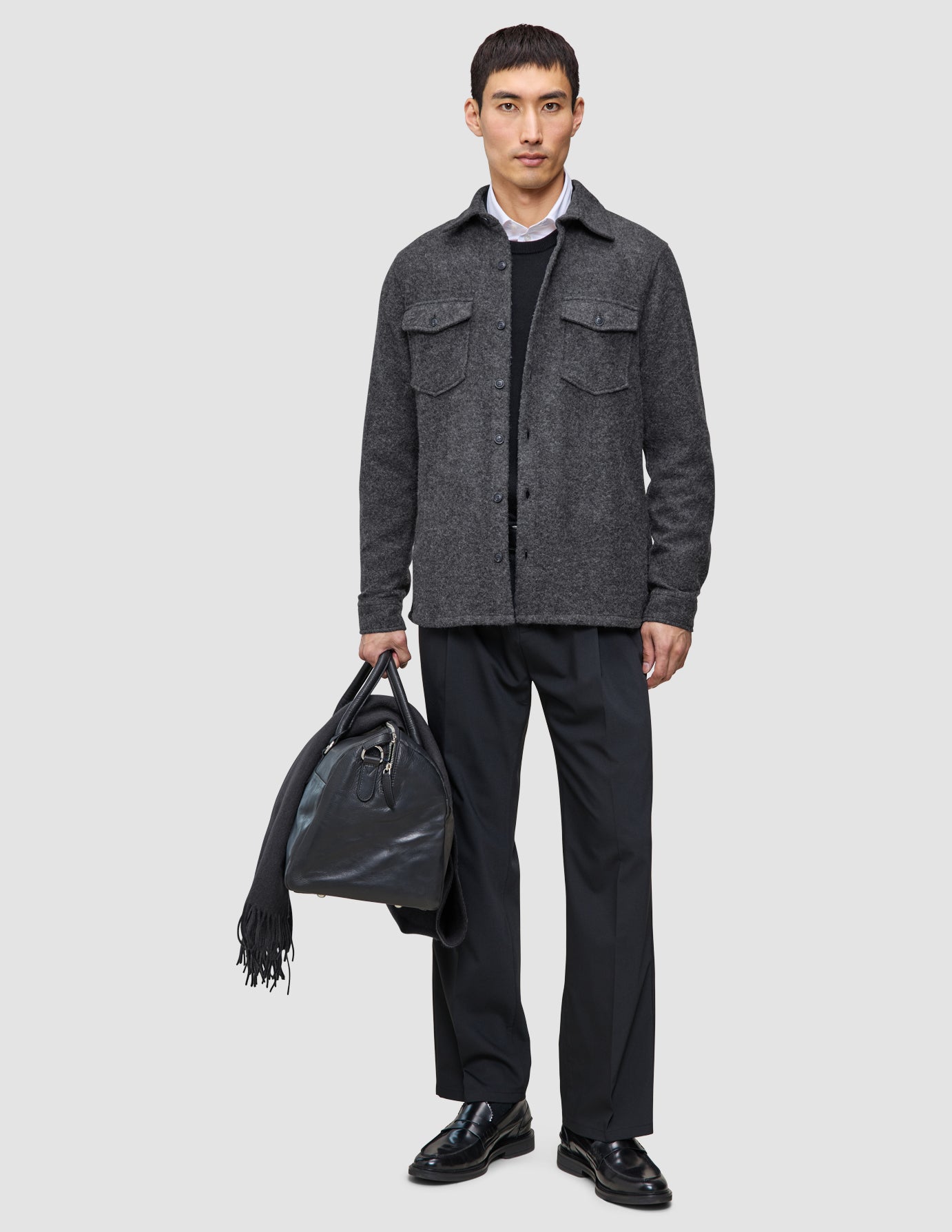 En man klädd i ett grått ylleöverskjorta, svarta funktionsbyxor och loafers, som håller i en svart läderresväska och en halsduk.