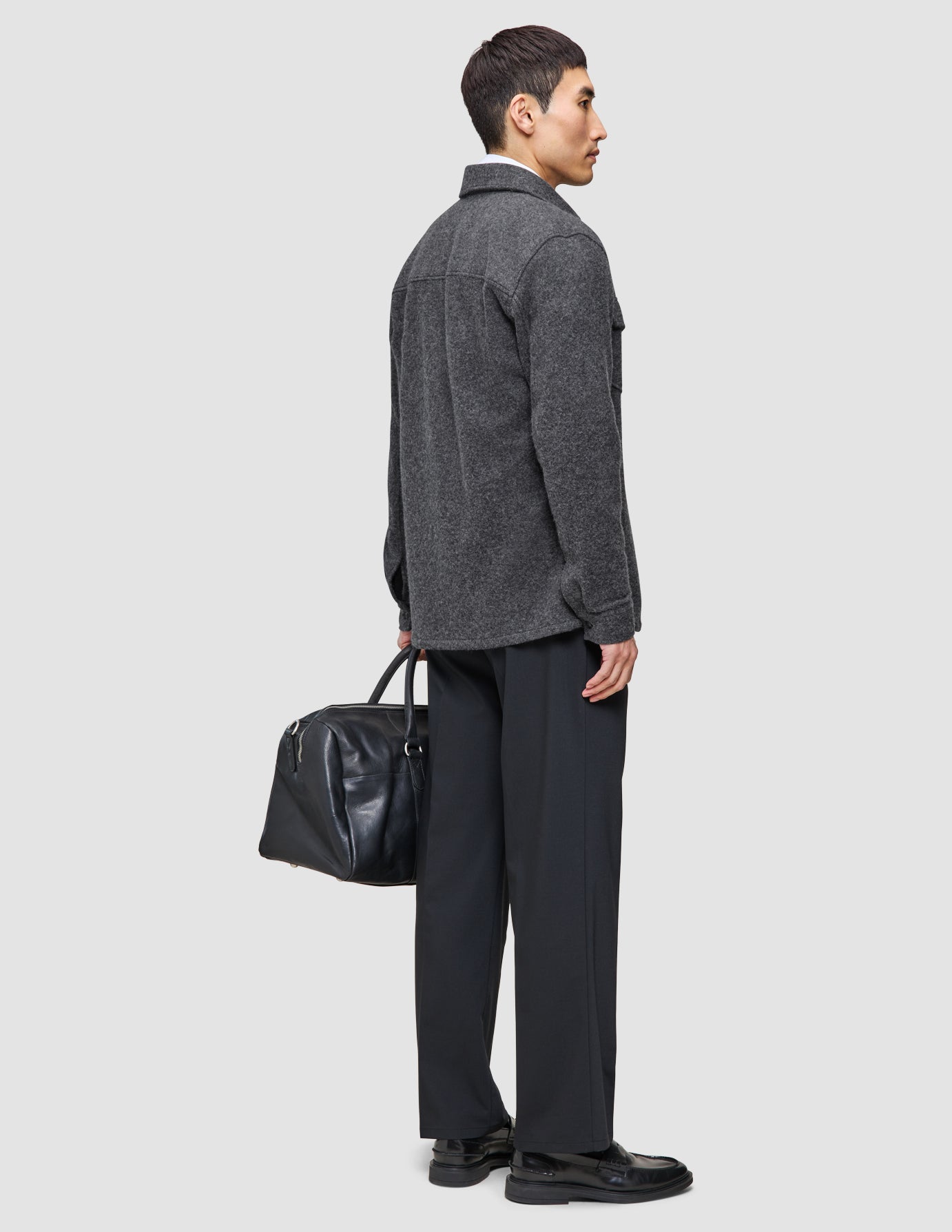 En man klädd i en grå dubbellagad ylleöverskjorta och svarta Tech Wool-byxor från Layers that work bär en svart läderväska.