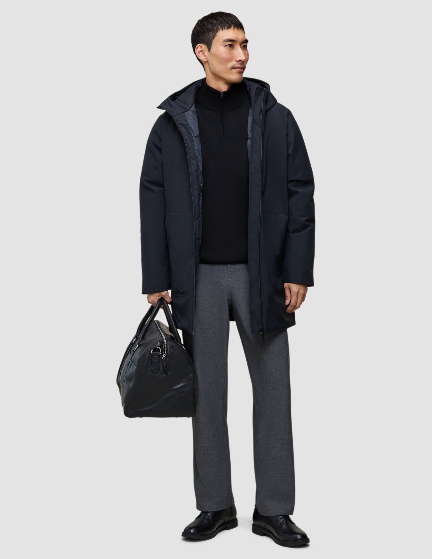 En man klädd i en svart Utility Jacket, en tröja och grå byxor bär en svart resväska.