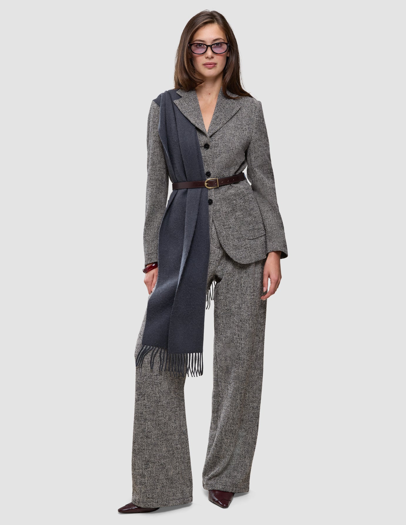 En person klädd i en grå blazer med fiskbensmönster och byxor av modellen Women Ashryn Pants Wide Domino Herringbone, med ett brunt bälte och en lång halsduk.
