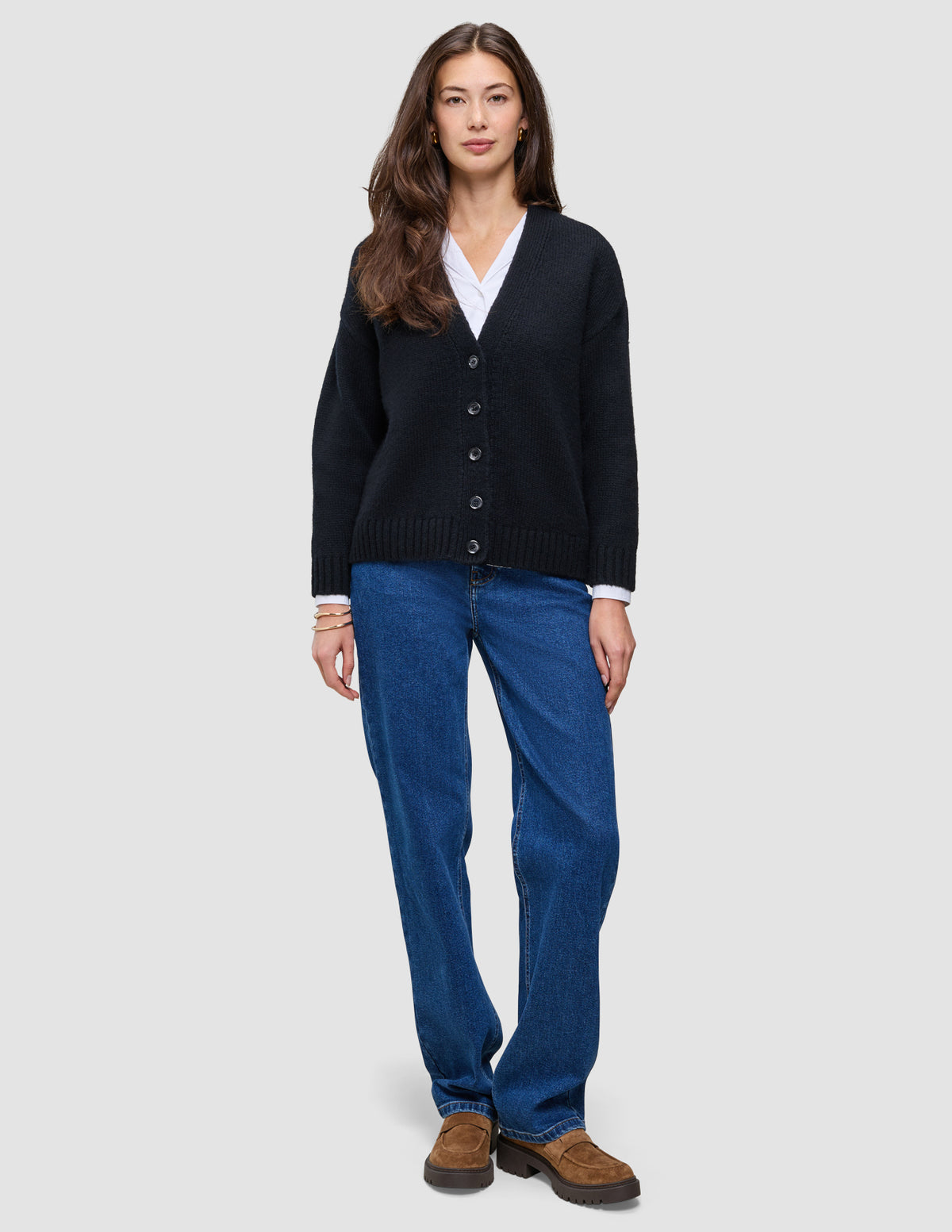 En person bär en svart, tjock och mjuk stickad damkofta i ull över en vit skjorta och blå jeans samt bruna loafers.