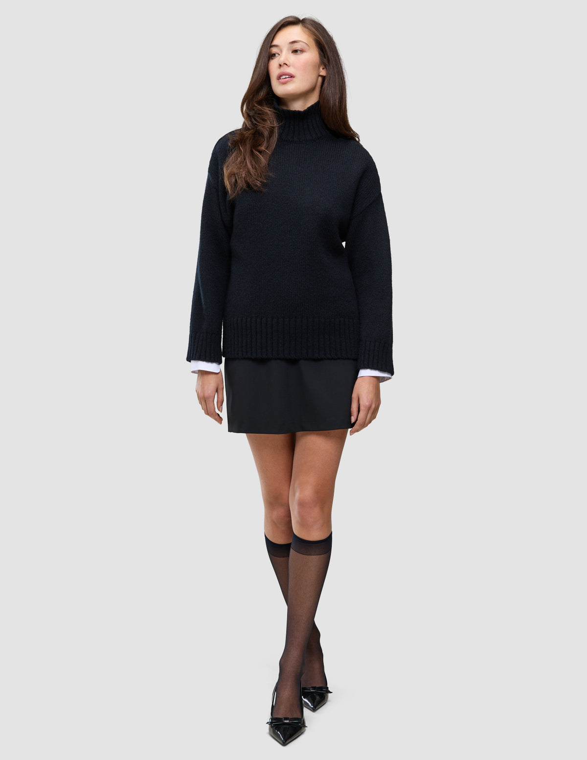 En kvinna i en tjock, mjuk, svart turtleneck i ull över en vit skjorta med krage, svart kjol, strumpbyxor och högklackade skor.
