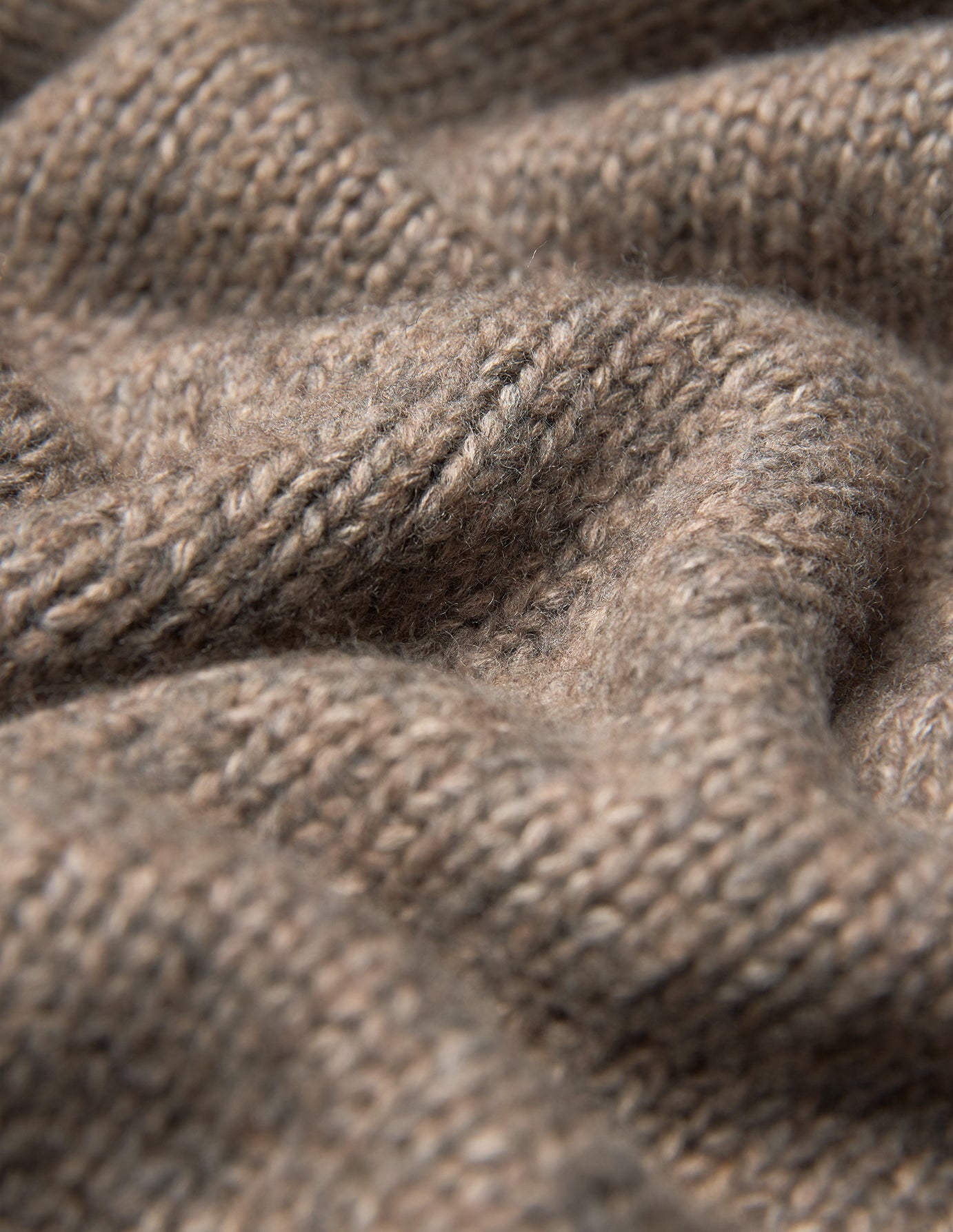 Närbild på en tjock, mjuk stickad damtröja med hög hals i taupe-melange, med en grov, ribbad struktur i taupe.