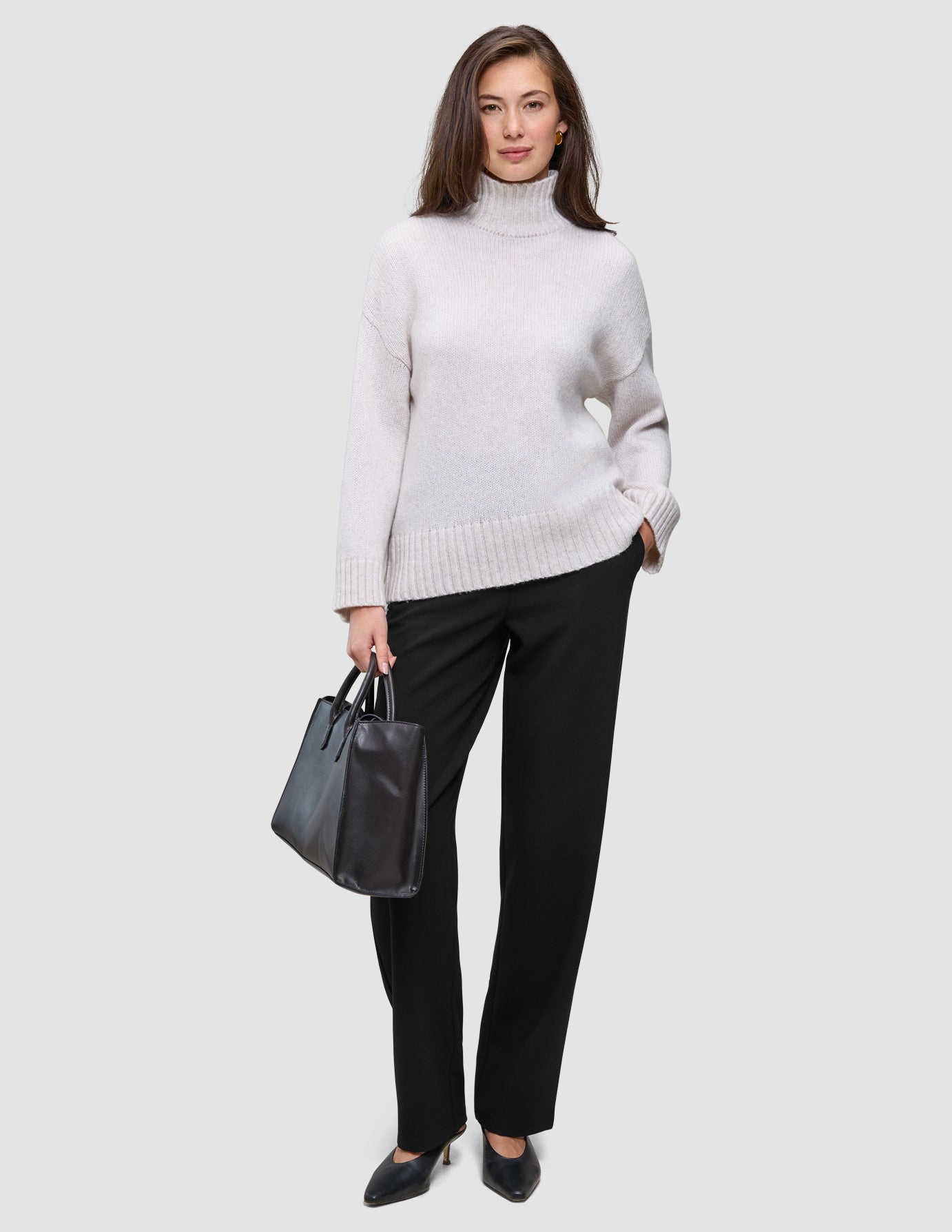 En kvinna klädd i en krämfärgad tröja med hög hals och svarta Women GO2 Pants Straight, som håller en svart handväska.
