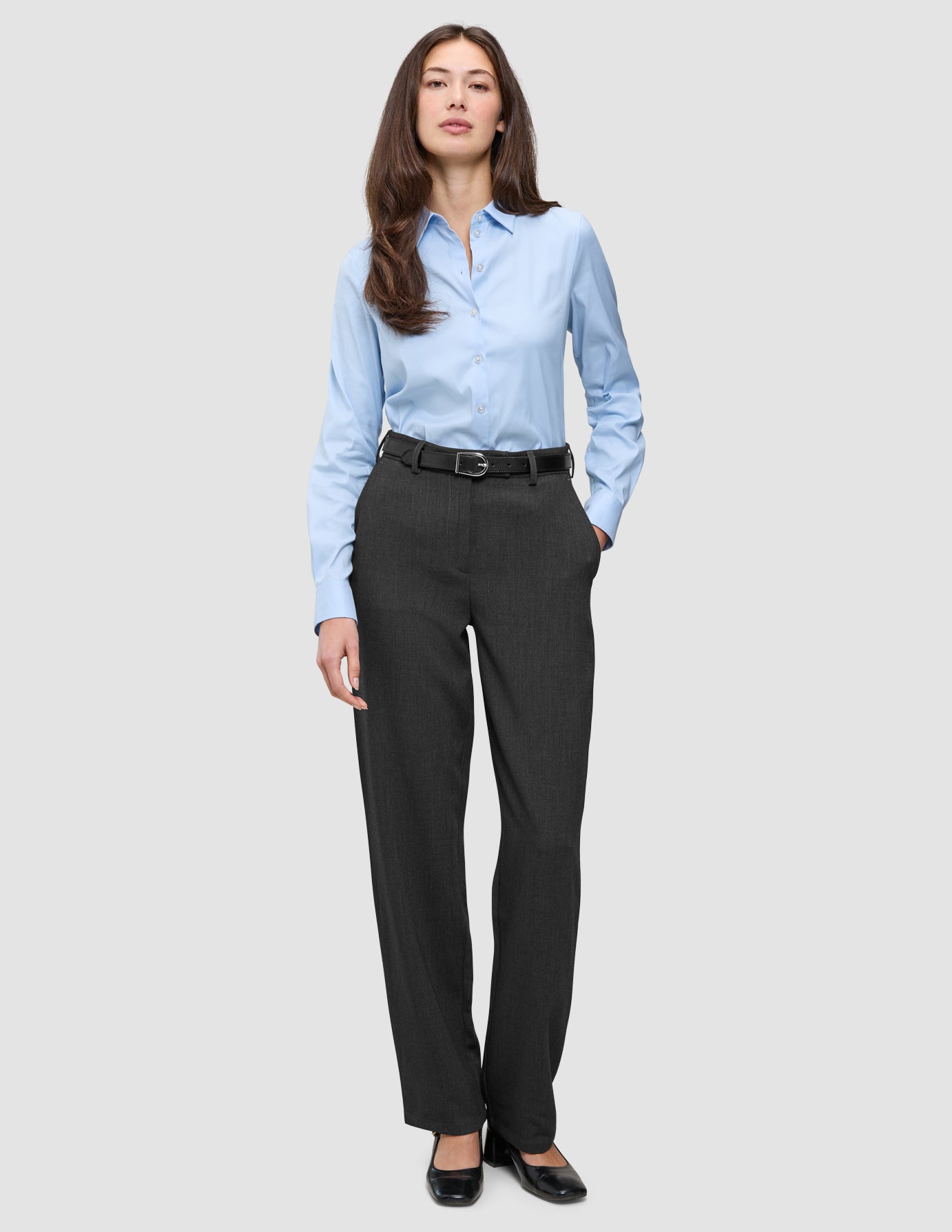 En kvinna klädd i en ljusblå skjorta med knappar, ett svart bälte och byxor av modellen Women GO2 Pants Straight i mörkgrå melange.