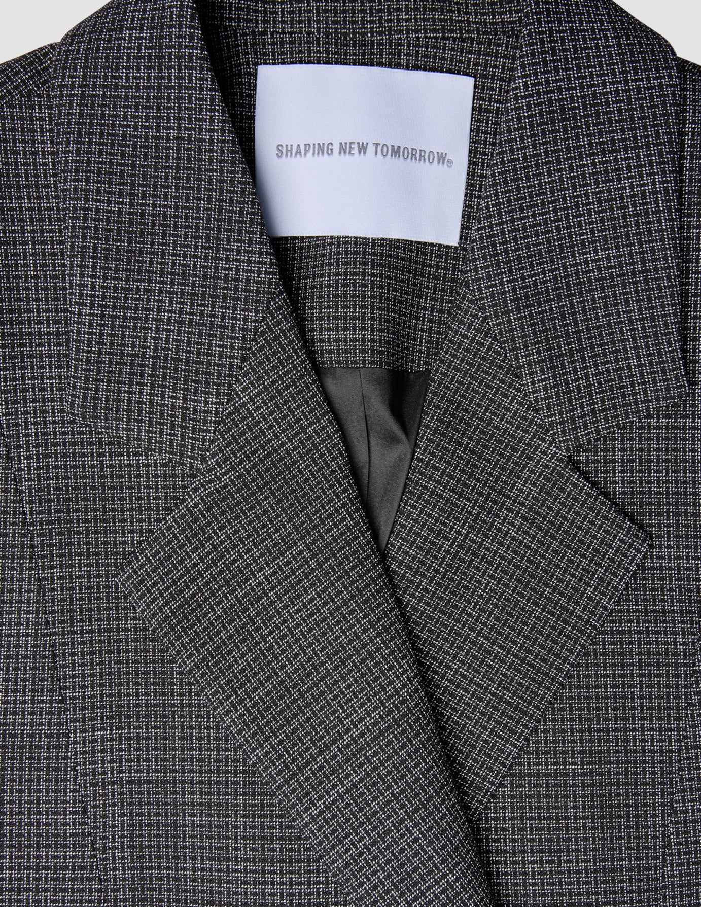 Närbild på en svartvit rutig damblazer av modellen Nara Blazer Fitted Black Check med en etikett i nacken med texten SHAPING NEW TOMORROW.