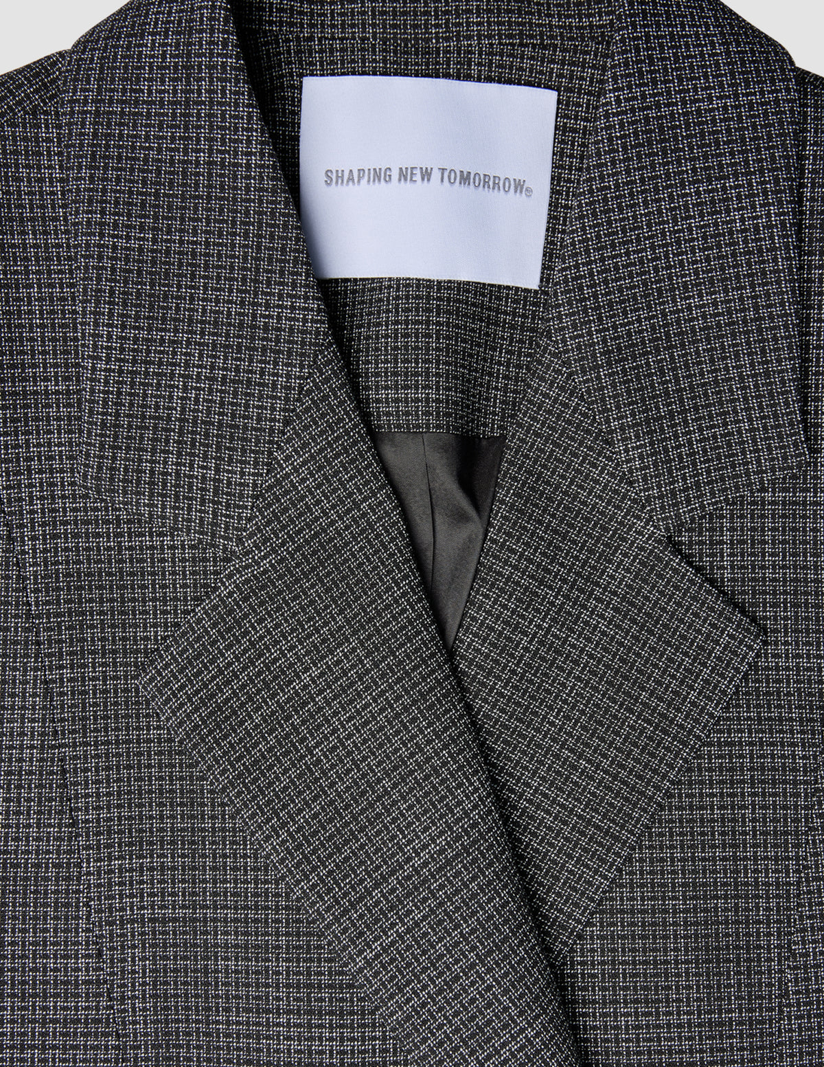 Närbild på en svartvit rutig damblazer av modellen Nara Blazer Fitted Black Check med en etikett i nacken med texten SHAPING NEW TOMORROW.