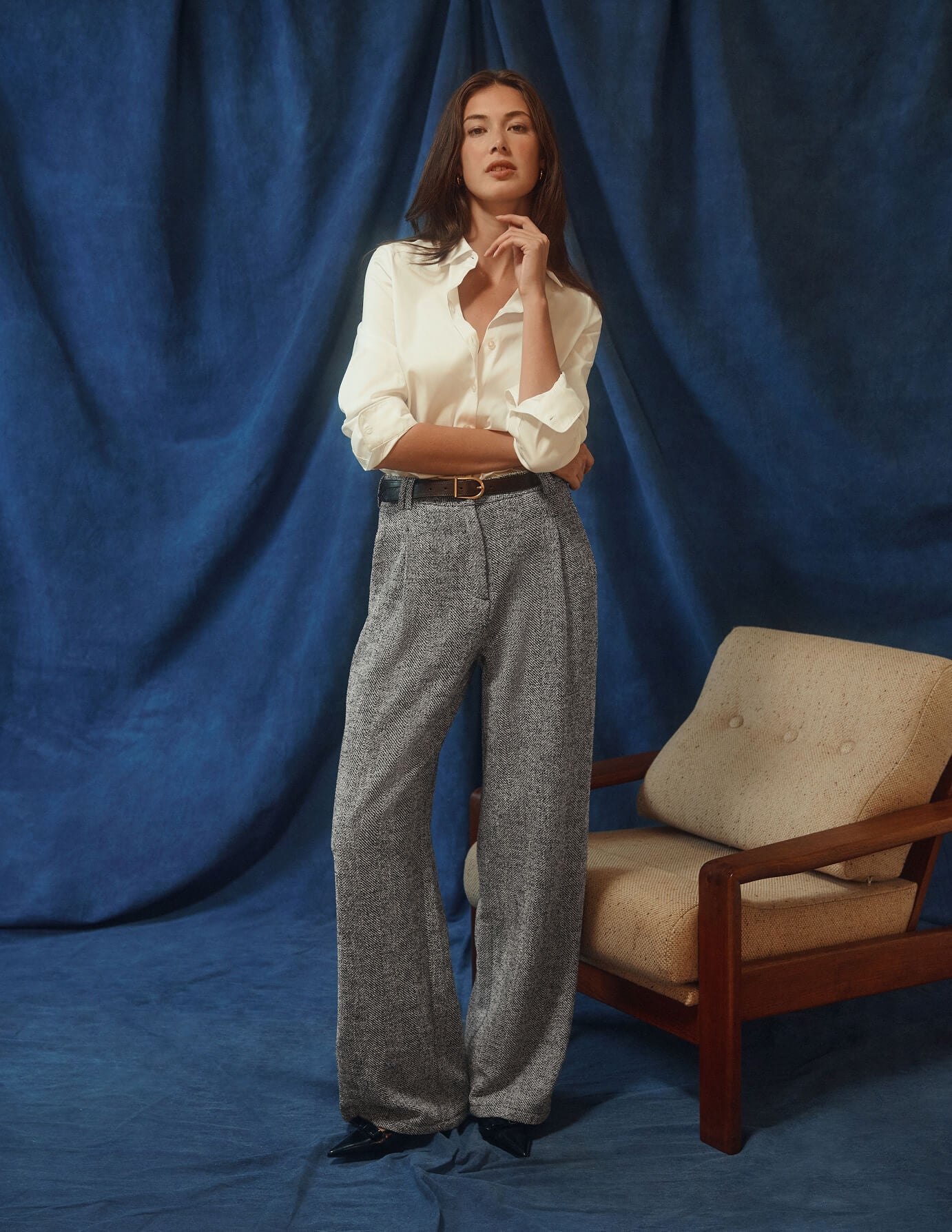 En kvinna klädd i en vit Women’s Oxford Shirt White Orchid Slim och grå byxor med vida ben står bredvid en fåtölj.