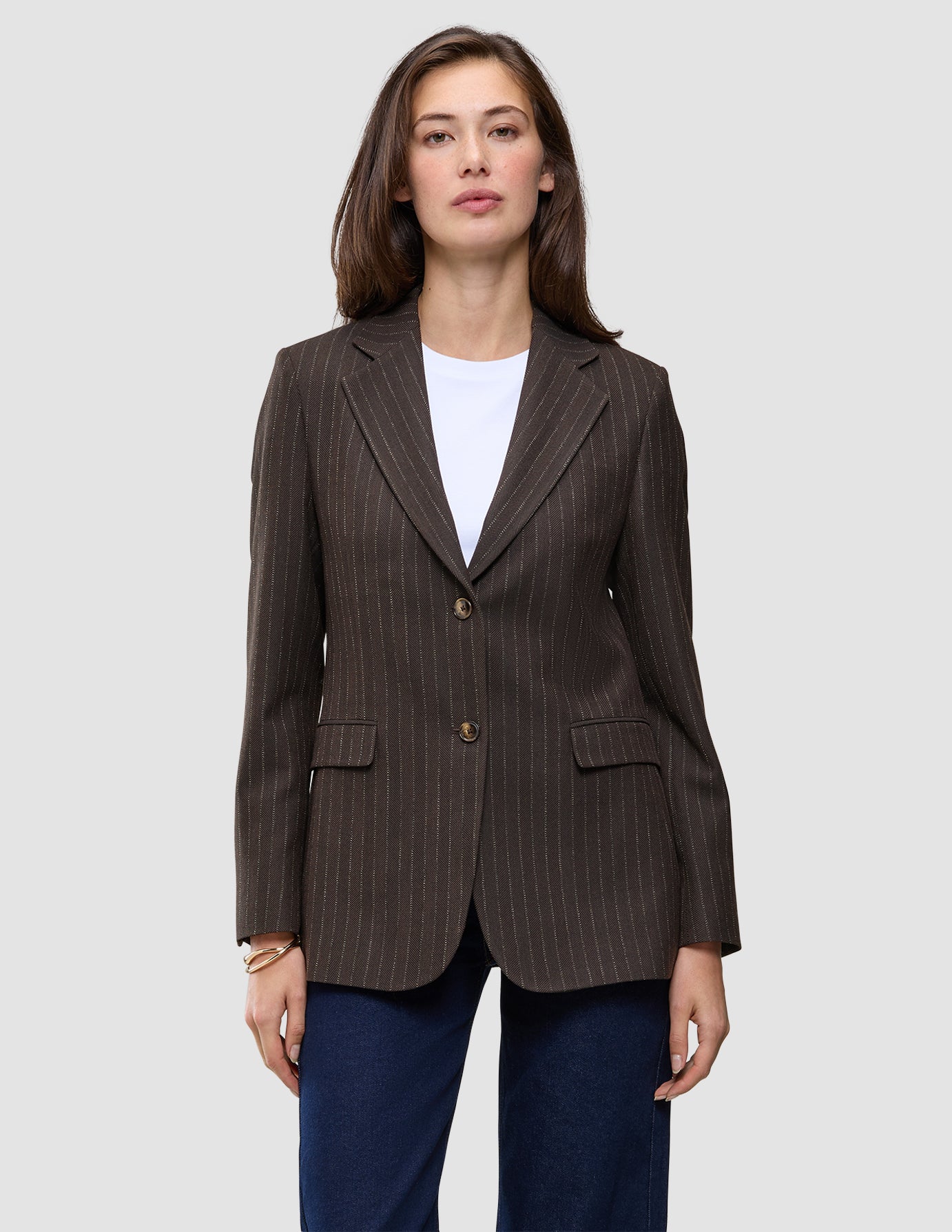 En kvinna bär en brun, valnötsfärgad Women Serene Blazer Regular i fiskbensmönster över en vit t-shirt och mörkblå jeans.