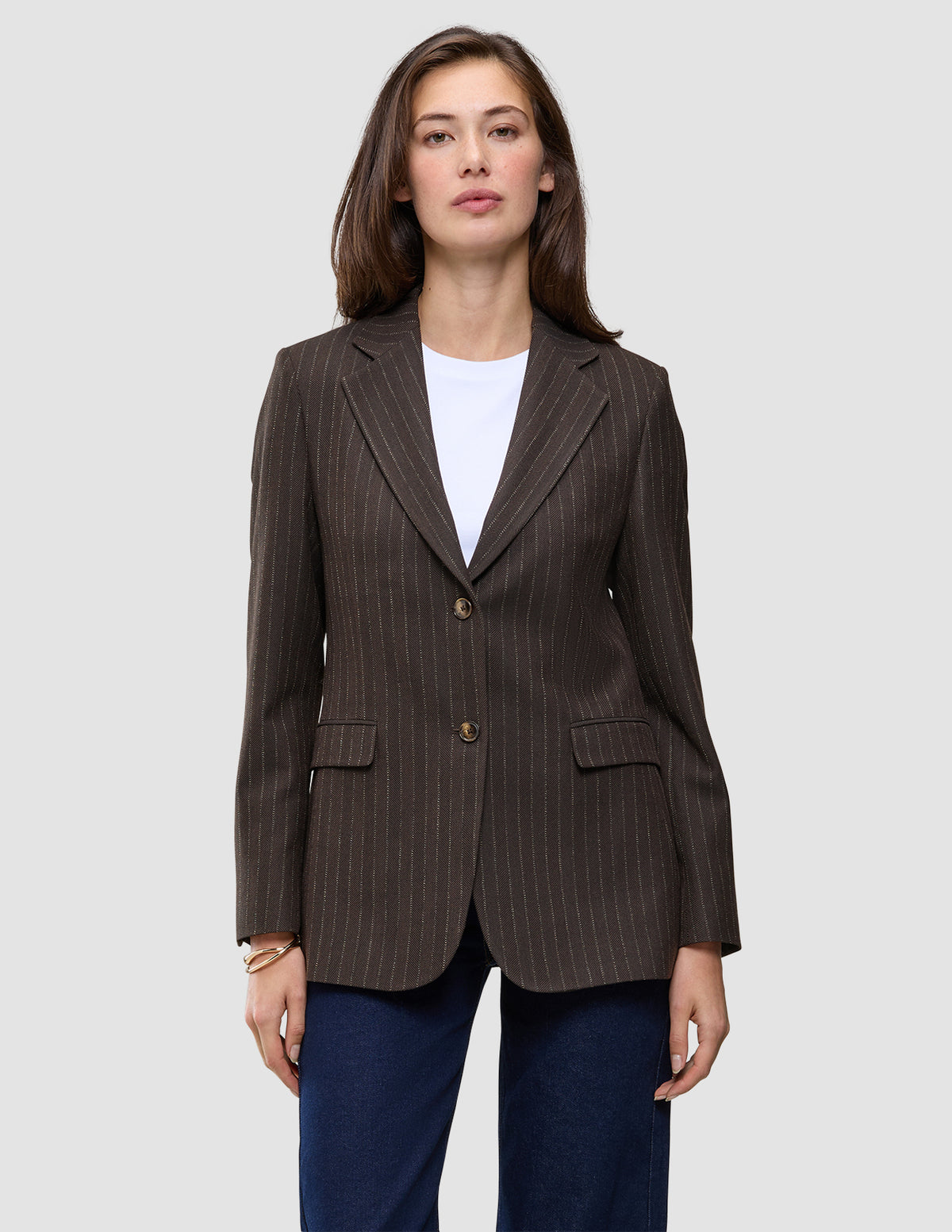 En kvinna bär en brun, valnötsfärgad Women Serene Blazer Regular i fiskbensmönster över en vit t-shirt och mörkblå jeans.