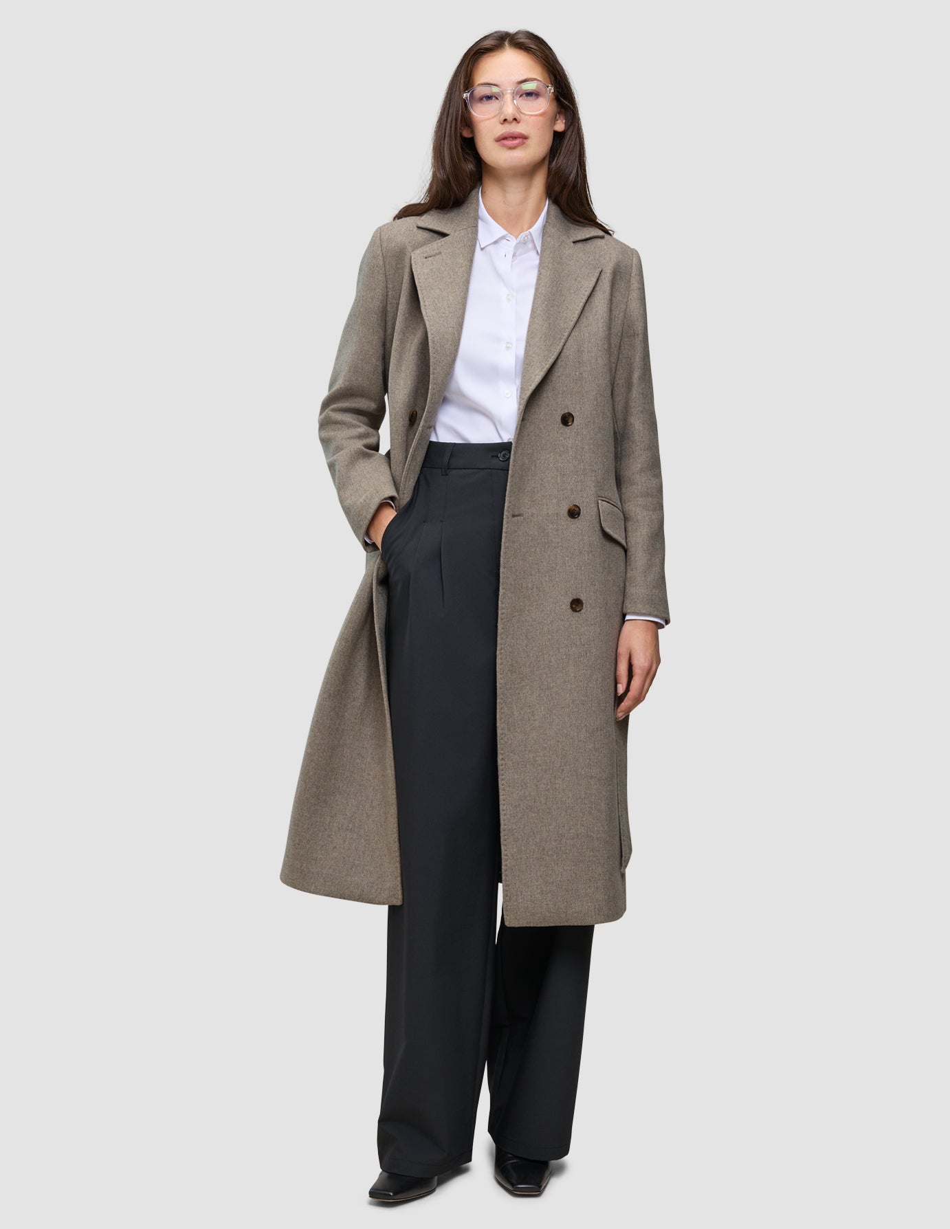 En person klädd i en lång, dubbelknäppt taupefärgad ullkappa i modell Women Wool Coat Greige över en vit skjorta och svarta byxor.