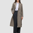En person klädd i en lång, dubbelknäppt taupefärgad ullkappa i modell Women Wool Coat Greige över en vit skjorta och svarta byxor.
