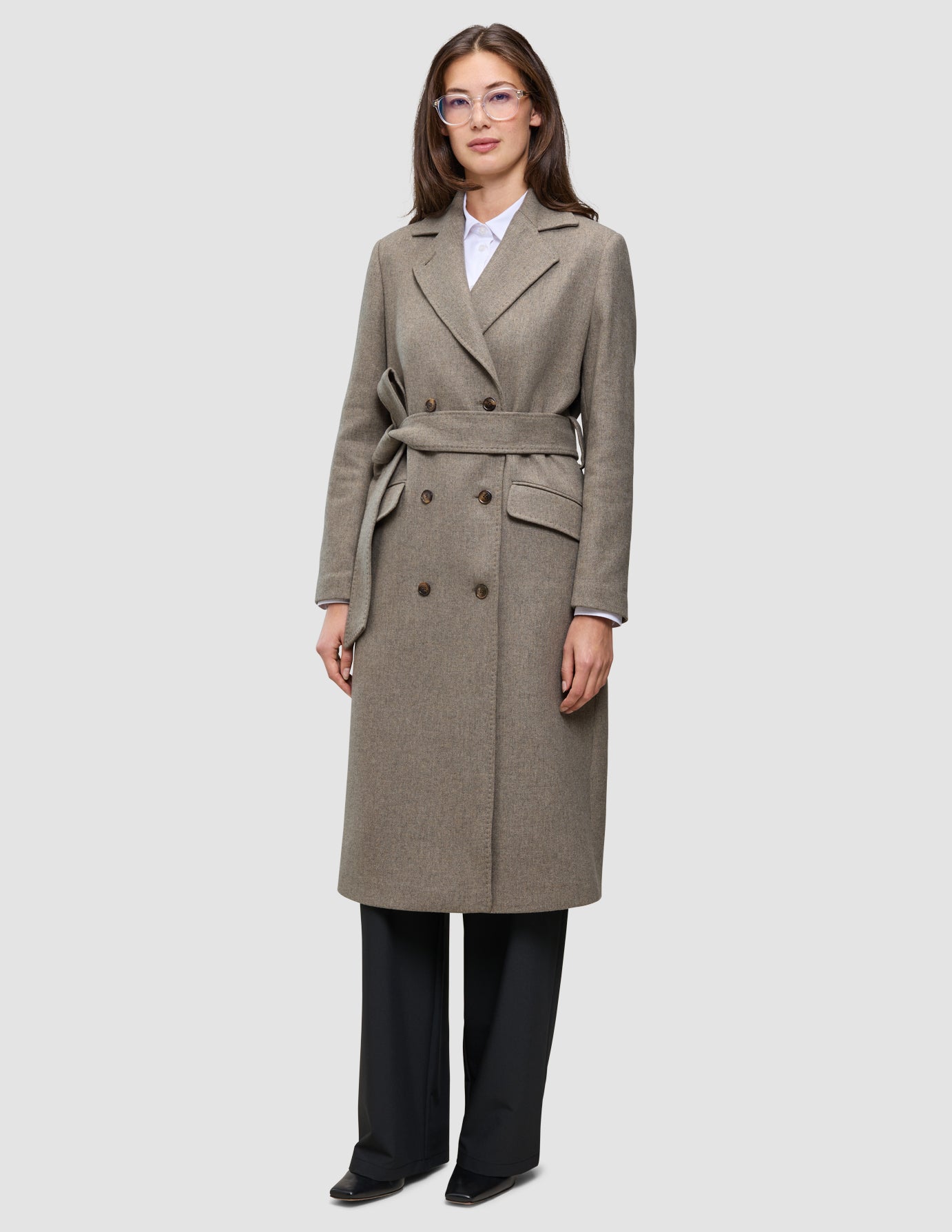 En kvinna i en gråbeige ullkappa för kvinnor, som hon bär över en vit skjorta och svarta byxor i skandinavisk design.